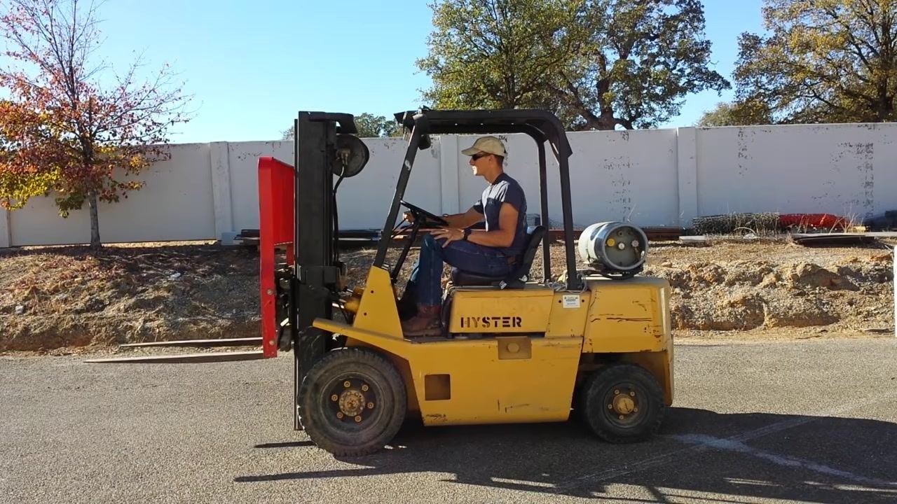 Side view of a yellow HYSTER forklift outdoors, with the operator seated upright, hands on the controls, wearing a cap and sunglasses. The forks are raised and slightly tilted, the propane tank is mounted at the rear, and the paved lot, a white wall, trees, and equipment are visible in the background.