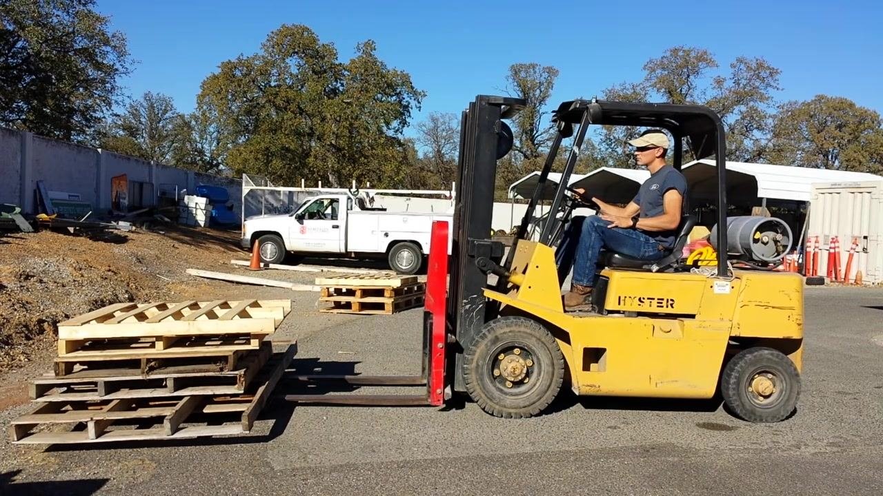 A yellow HYSTER forklift with a person operating it, forks positioned near a stack of pallets outdoors in a work yard.