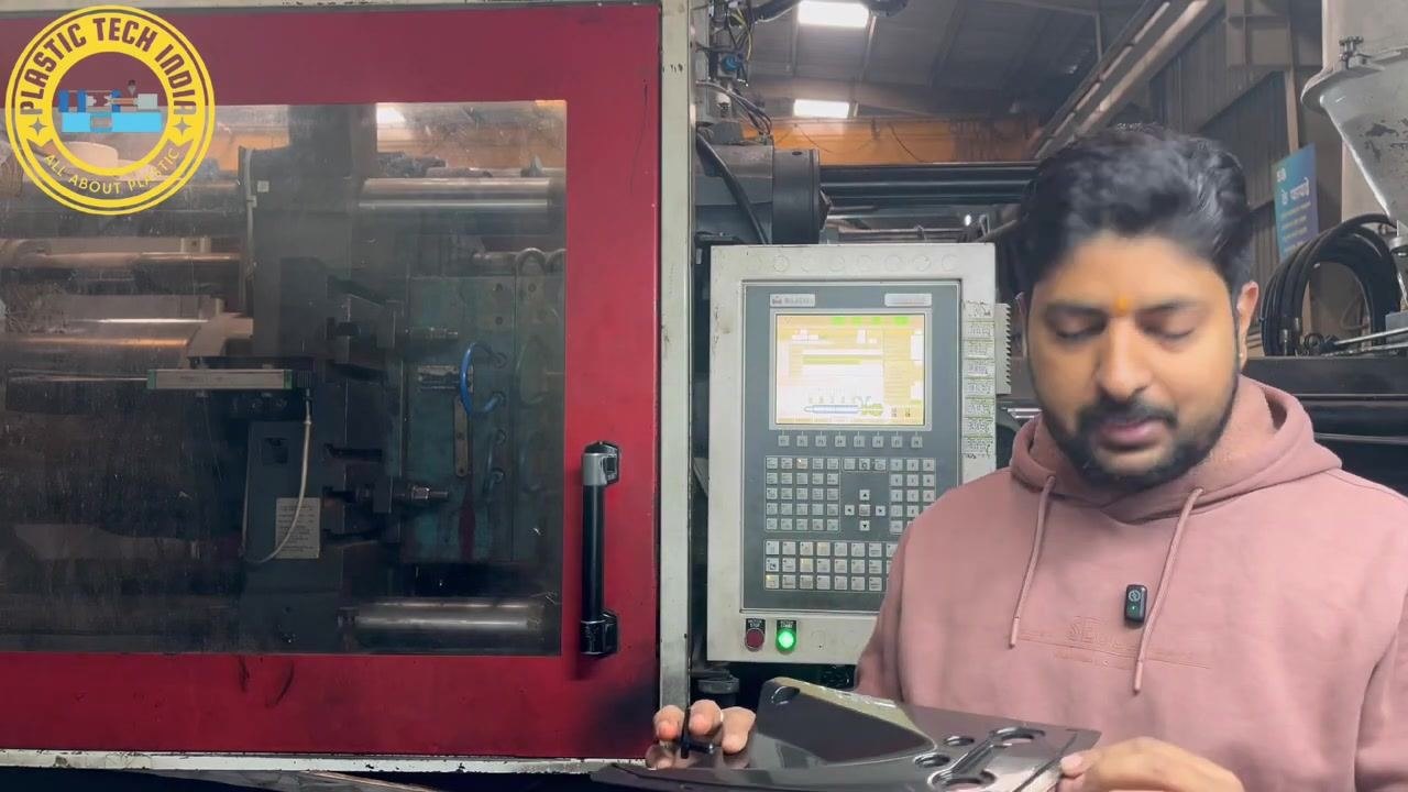 Presenter holding a rejected molded part that shows incomplete filling caused by the short mold defect, with the injection molding machine and control panel visible in the background.