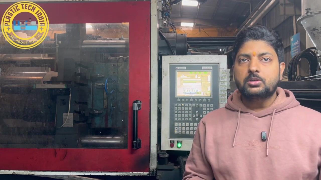 Presenter standing in front of the injection molding machine and control panel, emphasizing the importance of not making random adjustments.