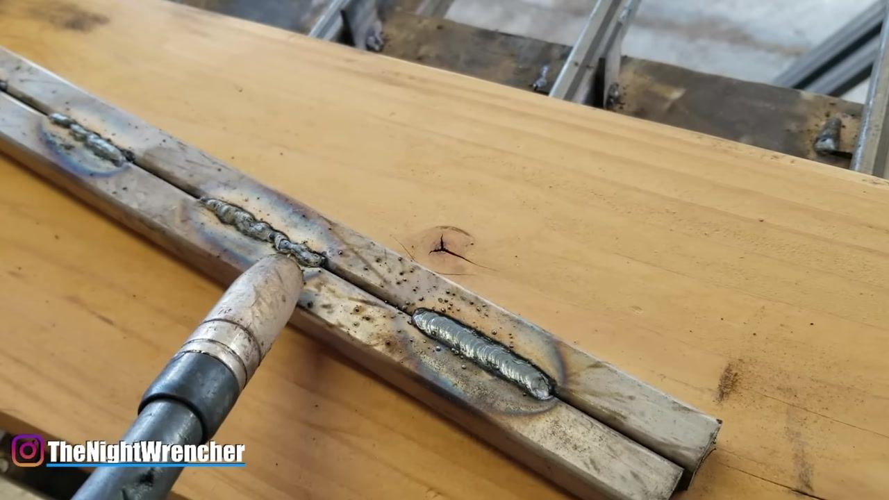A welding torch is positioned near several weld beads on two metal bars placed on a wooden workbench; the welds show varying quality and penetration.