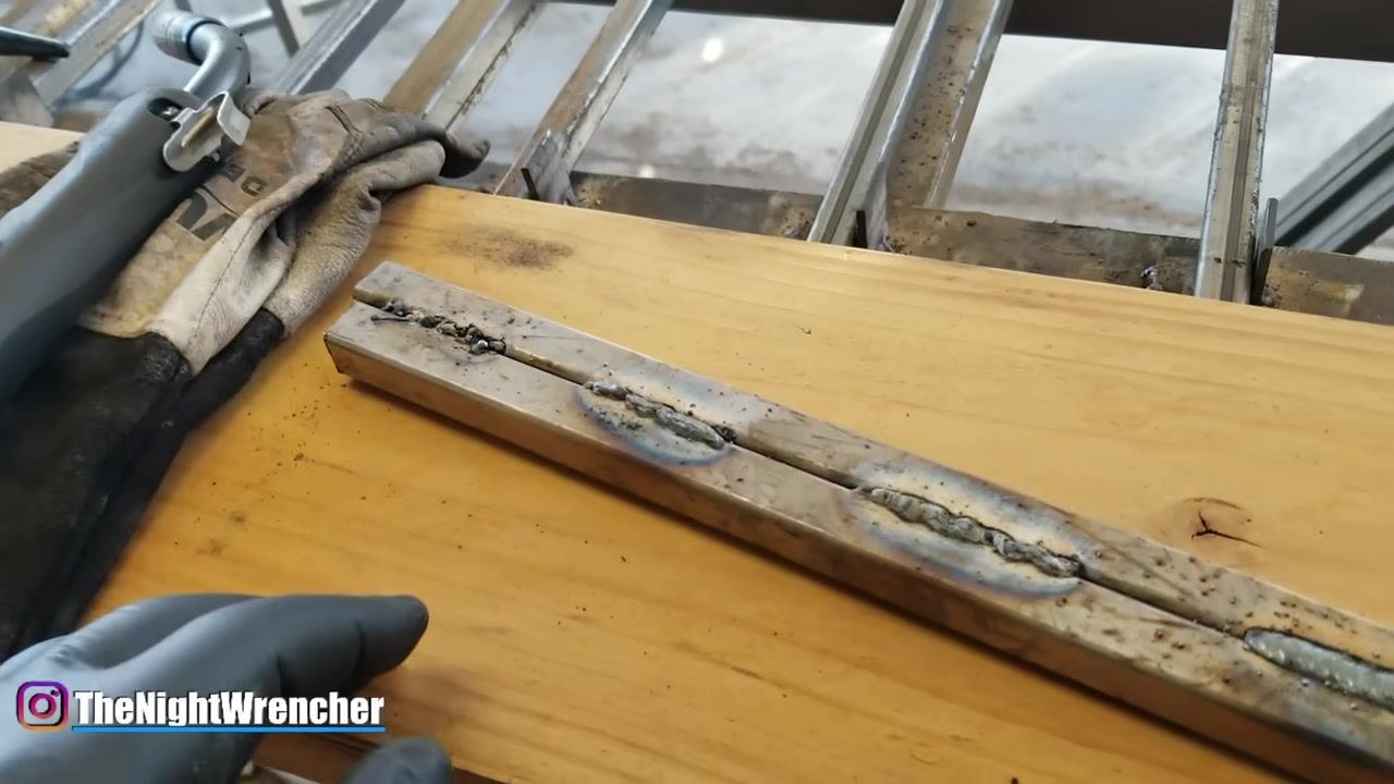 A gloved hand points at a weld bead on two metal bars placed on a wooden workbench, highlighting the importance of gun angle in welding technique.