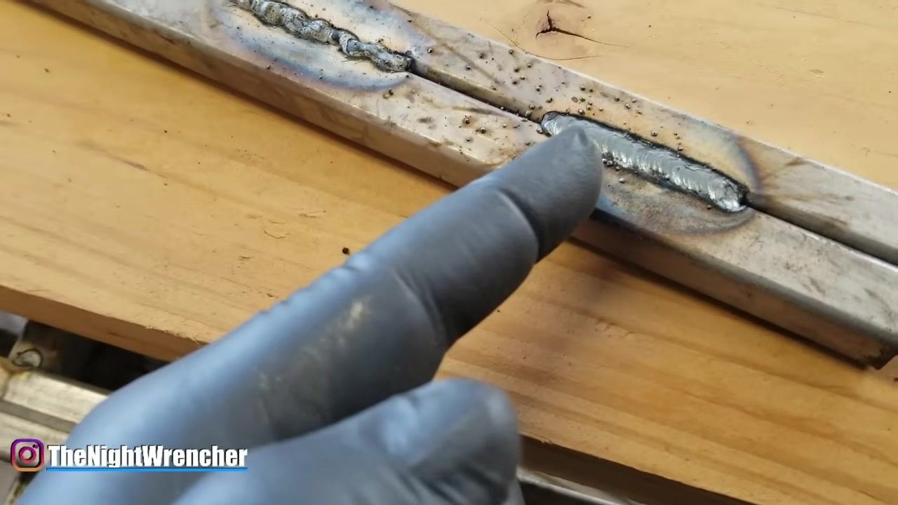 A gloved hand makes a V-shaped gesture above welded metal bars on a wooden workbench, illustrating the direction of weld puddle movement.