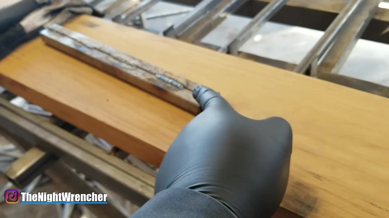 A gloved hand drags along the length of the welded seam on metal bars, demonstrating the proper motion for dragging the weld.
