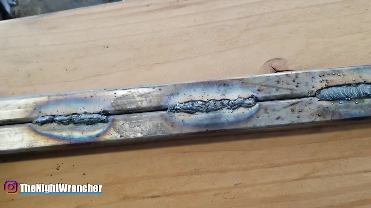 A gloved hand is positioned near two welded metal bars on a wooden workbench. The welds show visible slag and heat discoloration, demonstrating the result of dragging the weld puddle.