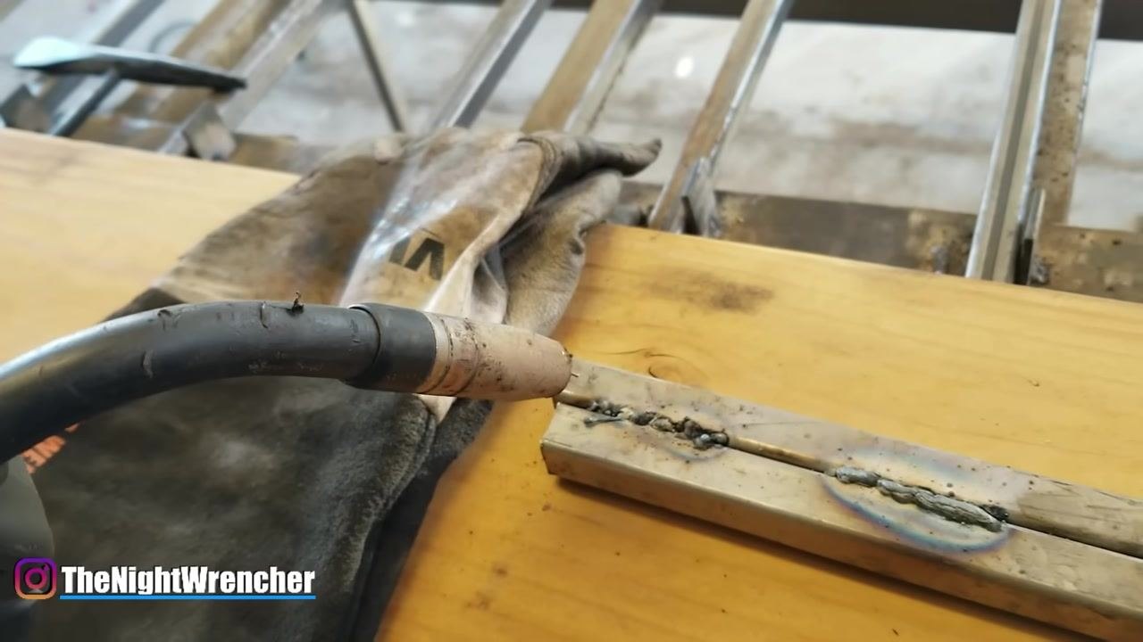 MIG gun positioned above a rough, spattered weld bead with visible globules; welding glove and metal sample on a wooden surface.