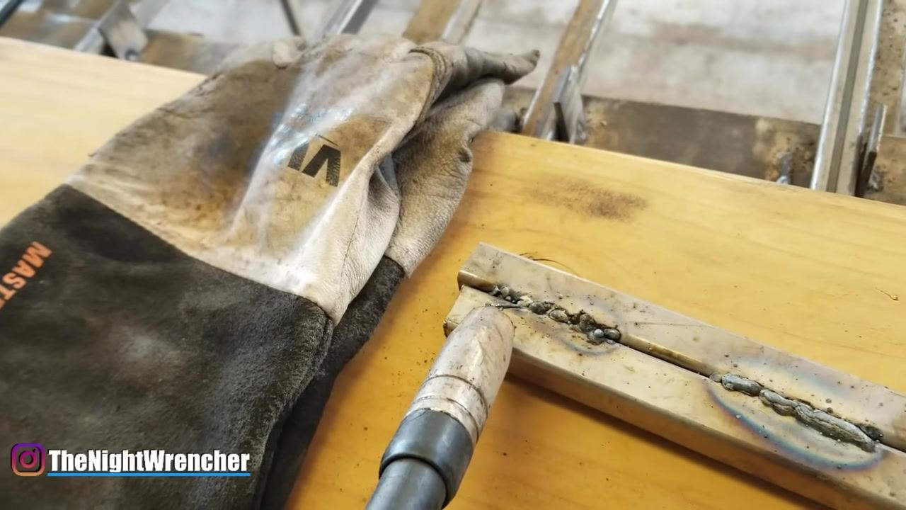MIG gun held above a weld bead with visible wire protruding, a rough weld surface, and spatter. Welding glove and metal sample on a wooden surface.