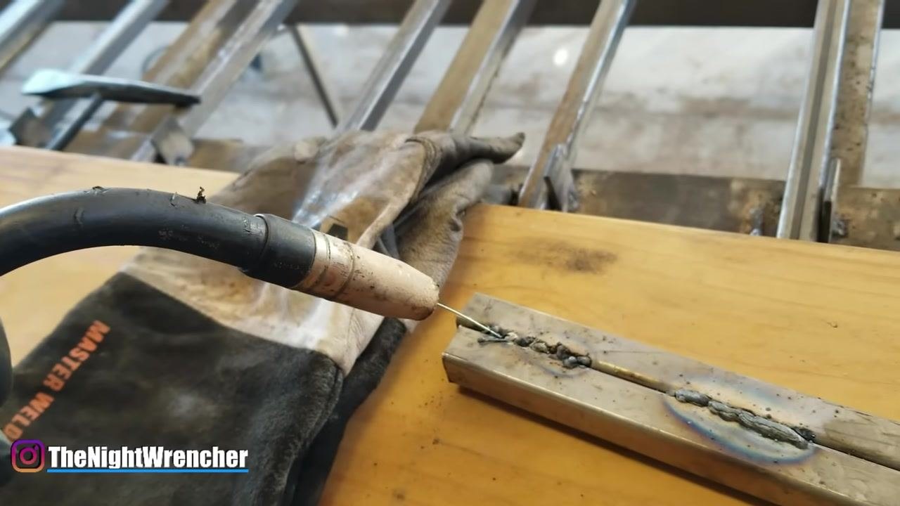 MIG gun held steadily above a more uniform weld bead with less visible spatter; welding glove and metal sample on a wooden surface.