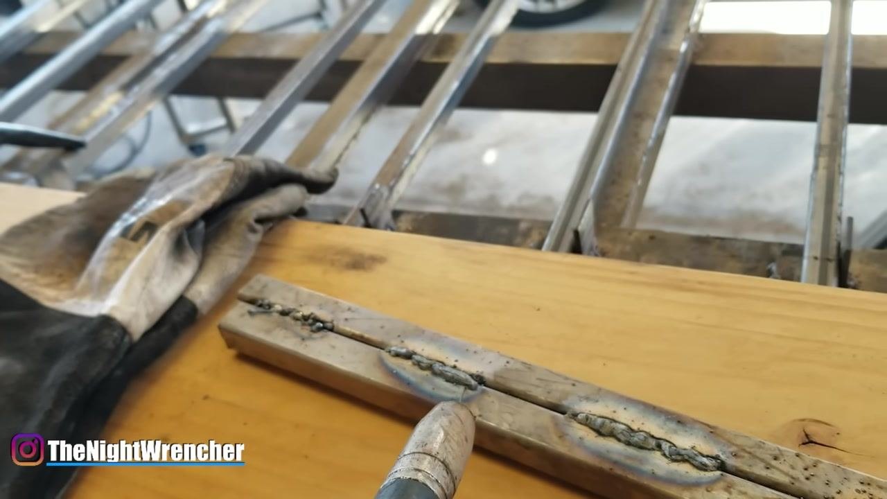 MIG gun positioned close to the metal workpiece, showing two weld beads and a welding glove on a wooden surface.