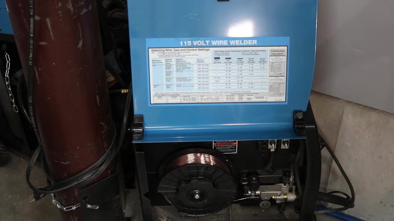 Close-up of a 115 Volt wire welder with the settings chart visible inside the open panel, showing wire, gas, and control settings for different material thicknesses and wire diameters