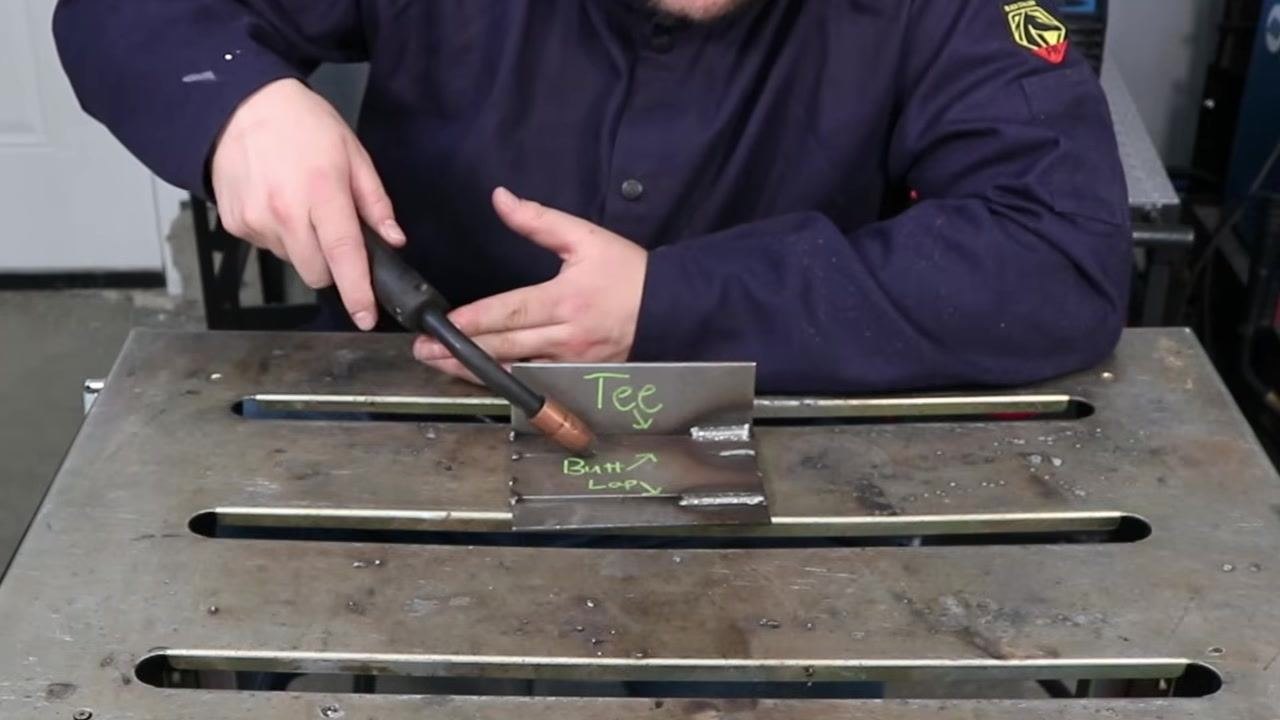 Person holding the MIG gun at an angle over the "Tee" and "Butt/Loop" plates, discussing angle importance in out-of-position welding