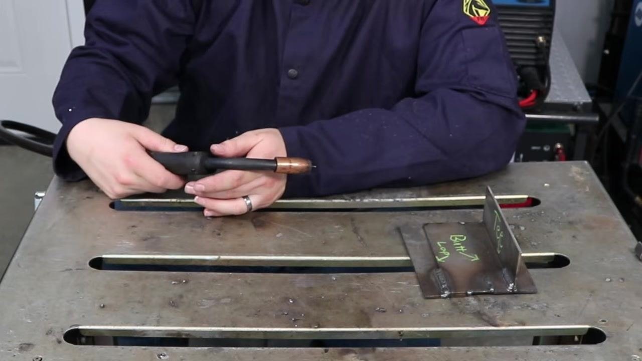 Person holding the MIG gun with both hands, workpiece set up for welding, emphasizing travel speed and movement