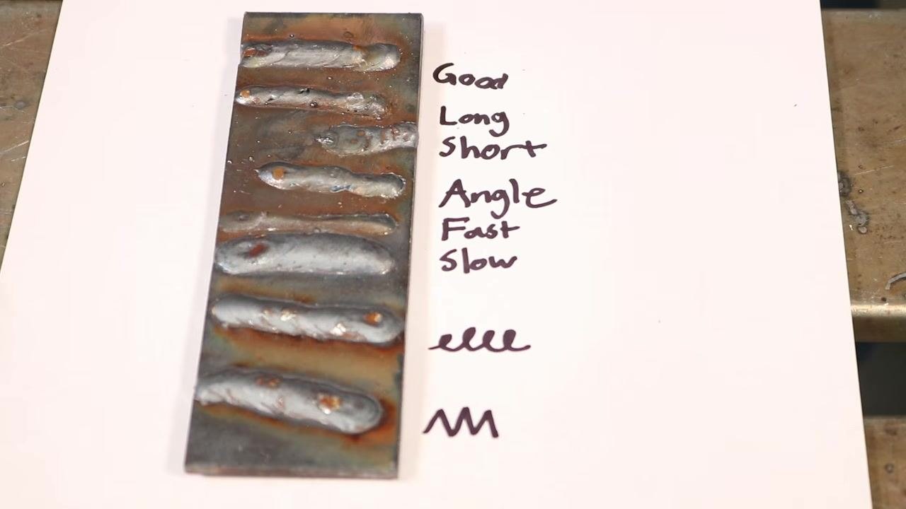 Person in a welding jacket holding a MIG gun above a metal workbench, with labeled metal pieces "Tee" and "Butt/Loop" nearby, demonstrating weaving technique options
