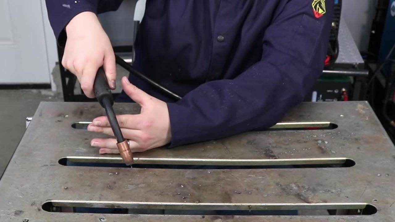 Welder demonstrating how to prop their hand vertically on the work table and use the other hand to guide the MIG gun, maintaining a steady stick out distance along the joint
