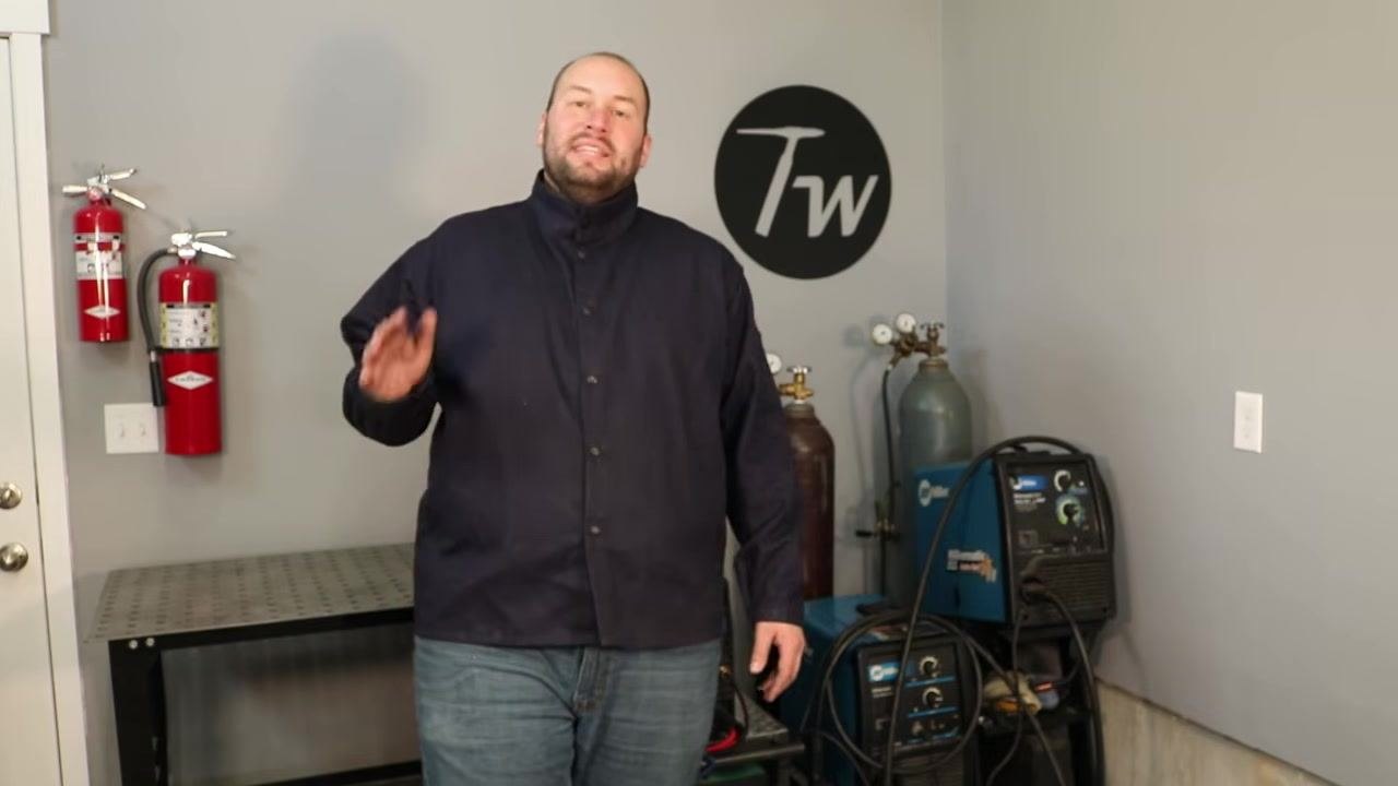 Instructor in a welding workshop, gesturing with hand, encouraging viewers to subscribe and continue learning