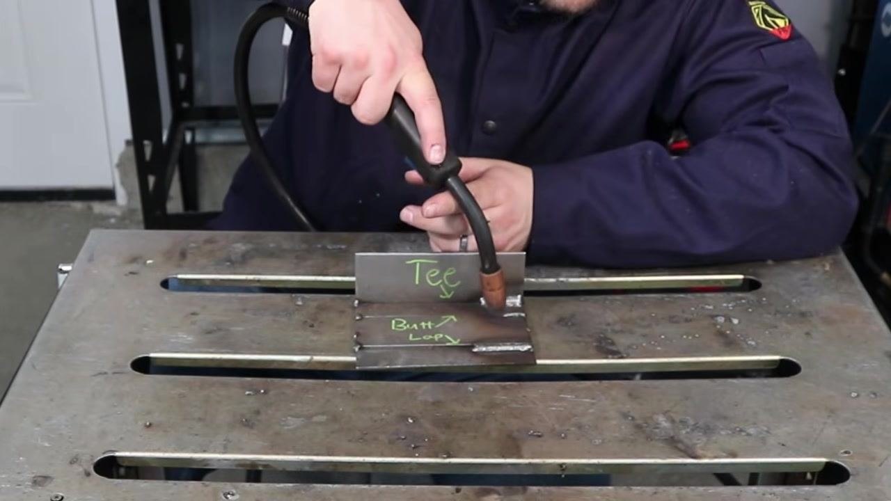 Welder demonstrating the correct work angle for a T-joint, holding the MIG gun at approximately 45 degrees to the joint, with labeled plates "Tee" and "Butt" visible on the work table