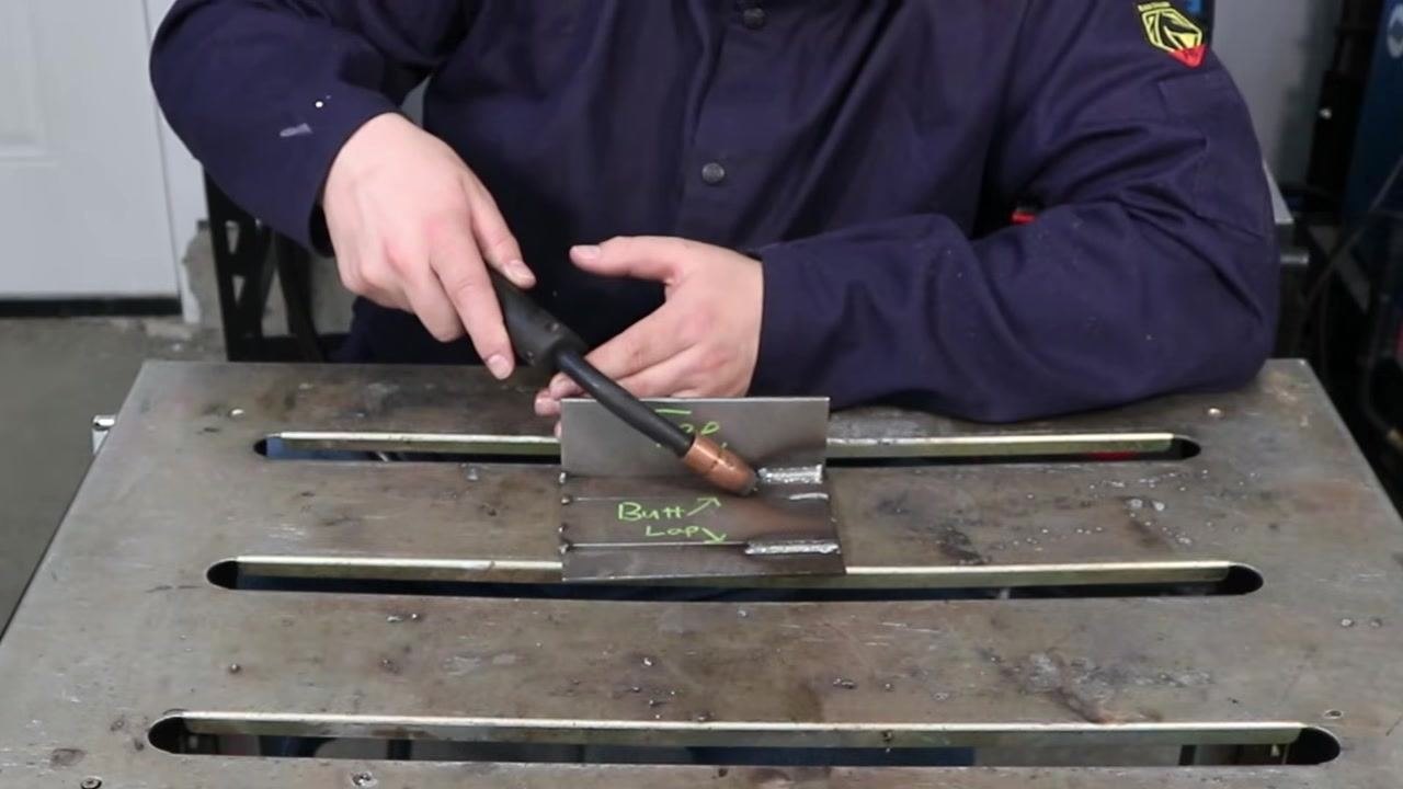 Welder demonstrating the MIG gun at the end of a dry run, showing consistent angle across the T-joint with labeled plates "Tee" and "Butt" visible