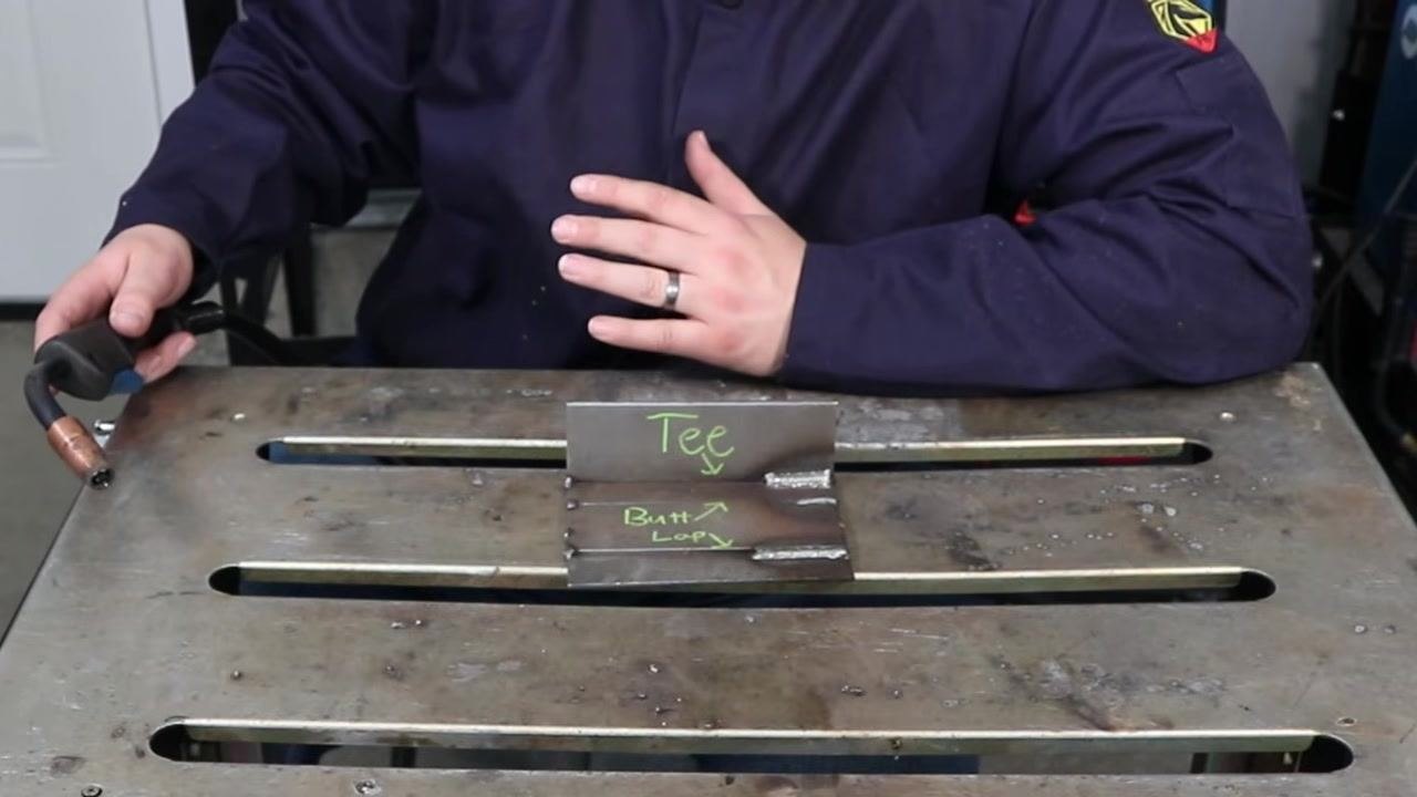 Person in a welding jacket holding a MIG gun above two labeled metal plates, "Tee" and "Butt/Loop," on a metal workbench, demonstrating dry run practice