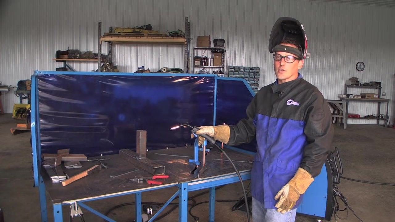 Welder standing at the workstation, helmet lifted, holding the MIG gun and addressing the camera, with a well-equipped shop in the background
