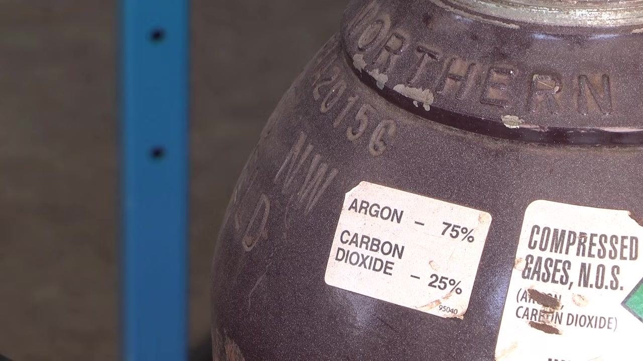 Close-up of a gas cylinder with regulator, labeled Argon 75 percent and Carbon Dioxide 25 percent, with a welding helmet on the table