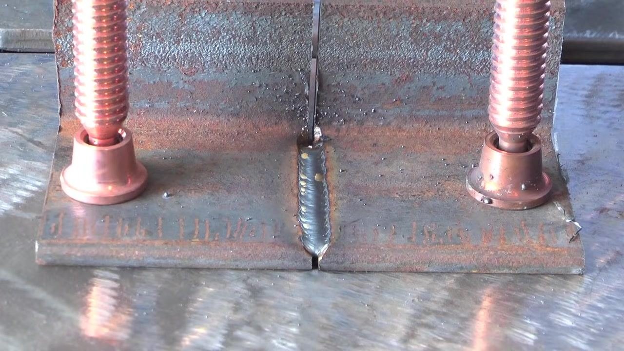 Close-up of a completed T-joint weld, showing clamps holding two metal plates at a right angle with a visible weld bead in the joint