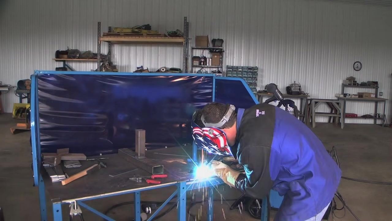 Welder wearing gloves and a helmet with an American flag design, positioning the MIG gun for an overhead weld on a steel structure