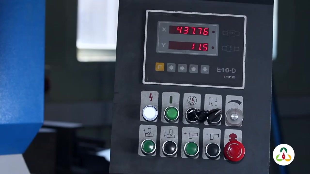 Operator wearing safety glasses and gloves, holding a metal plate and referencing a paper template to check the radius shape. Safety signage and "40X2000" machine label visible in the background.