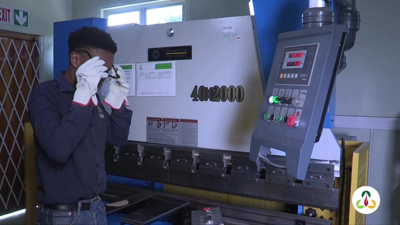 Close-up of gloved hands holding the metal plate in position on the press brake bed. Control box visible.