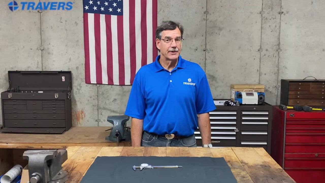 Instructor in blue TRAVERS shirt standing behind a workbench with a caliper on a mat, workshop background with toolboxes and American flag.