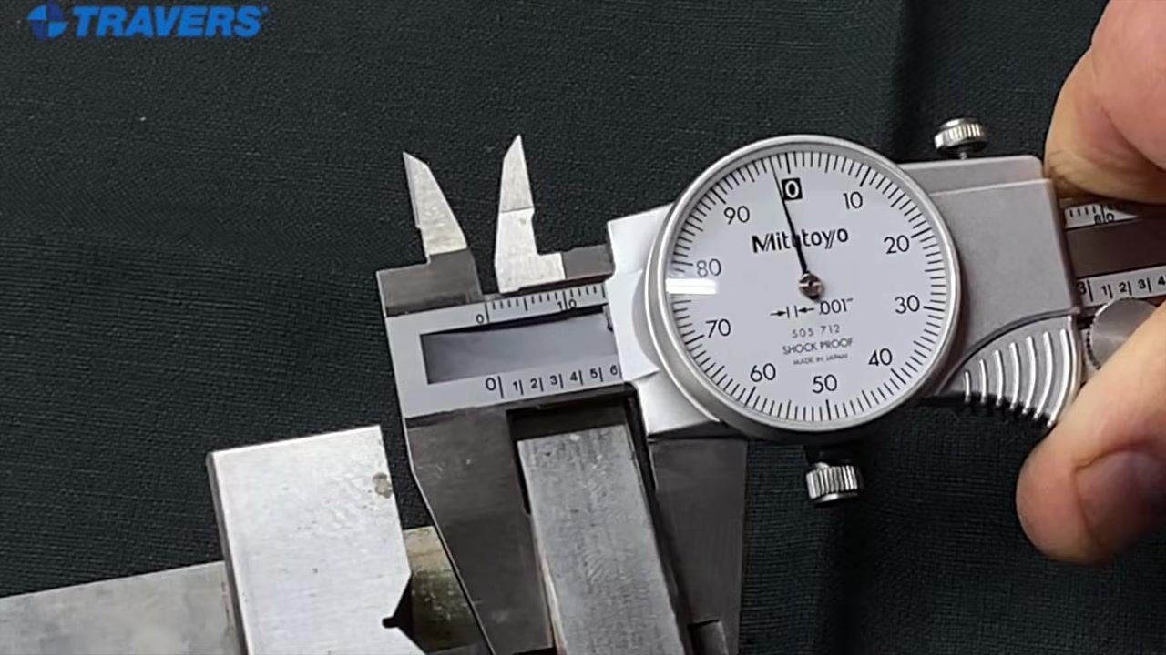A hand holding a Mitutoyo dial caliper with the thumb on the adjustment wheel and the jaws positioned around a metal object.