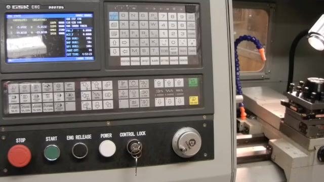 Operator using the MPG hand wheel on the CNC controller to manually move the tool carriage, with the controller display, MPG selector, and tool post visible.