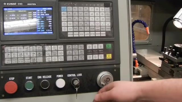 CNC controller with an operator's hand on the MPG hand wheel, showing the display, axis selection, and increment buttons, with the tool post and work area visible in the background.
