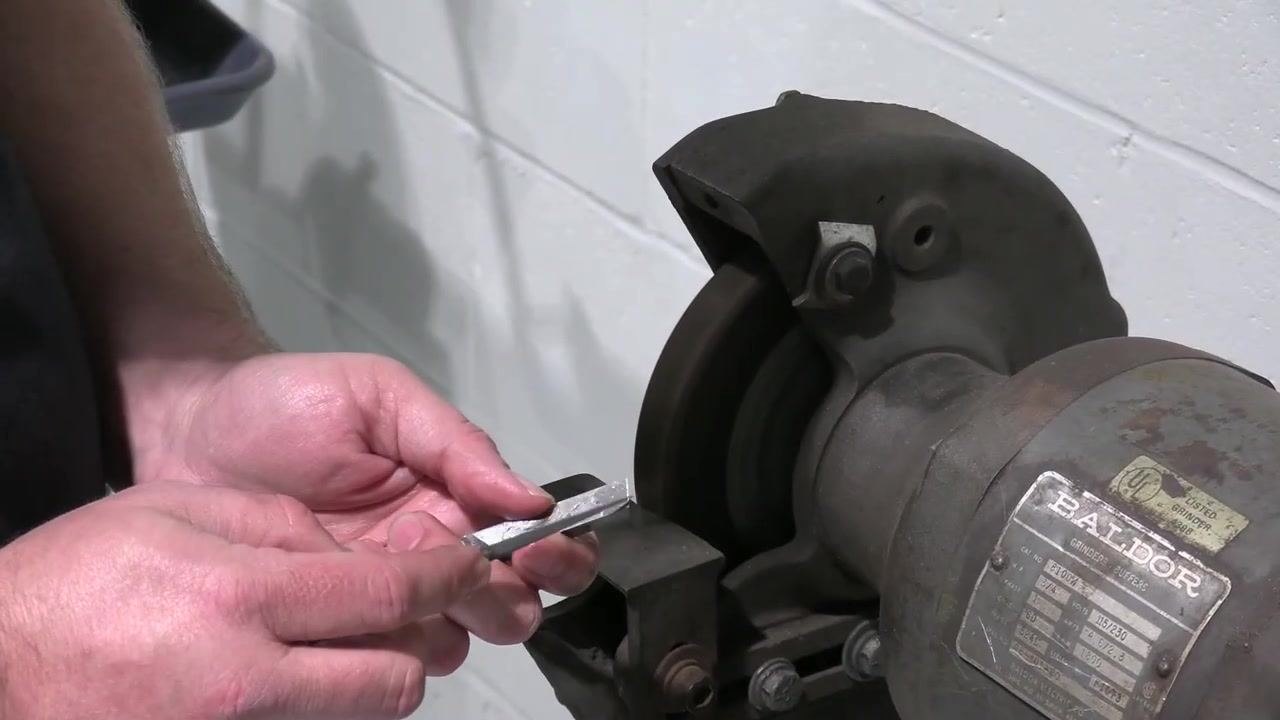 The finished tool bit is held in front of the bench grinder, showing the ground surfaces and overall profile.