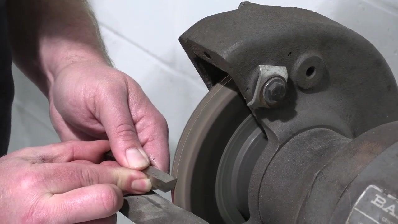 The tool bit is held in both hands for a final inspection before the test cut, with the bench grinder visible in the background.