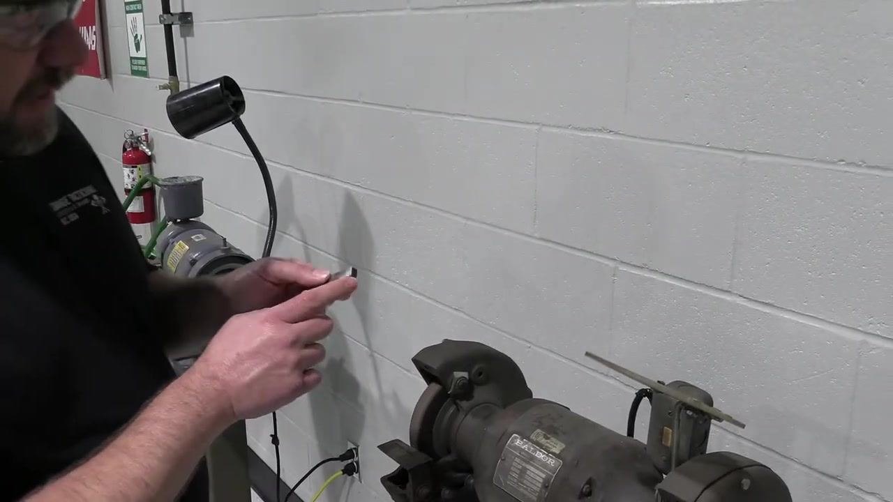 A hand points to the slanted side of the tool bit near the bench grinder, with safety signage visible in the background.