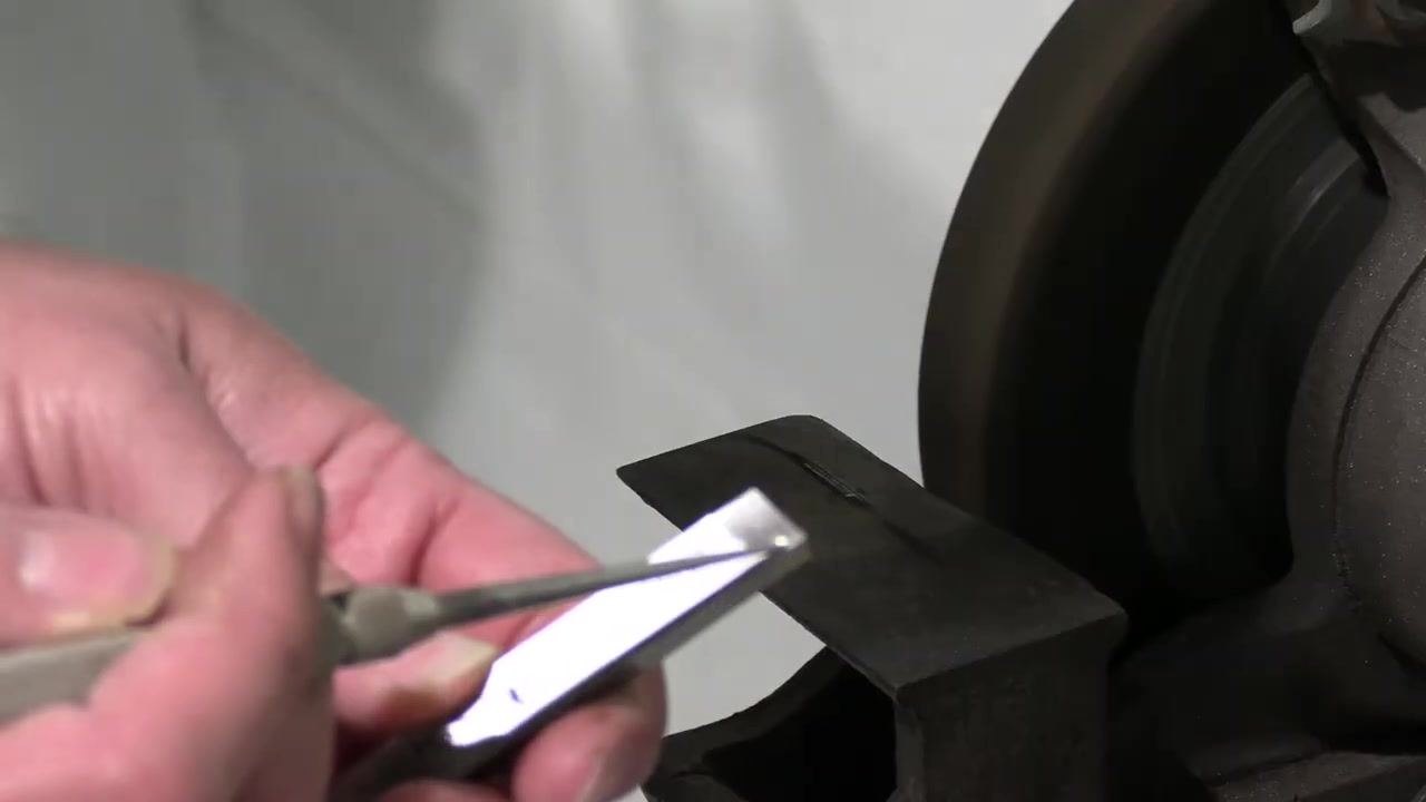 The tool bit is held under a light to inspect the reflection for uniformity, with the grinding wheel and tool rest visible.