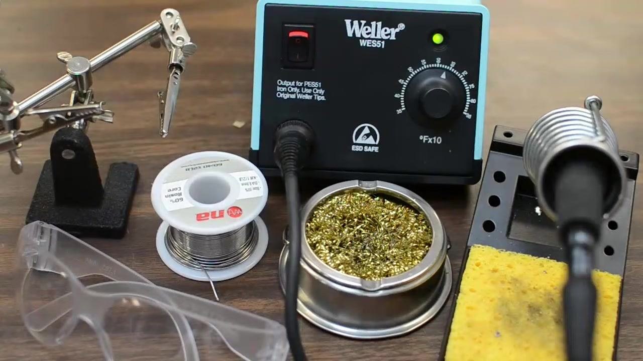 A complete soldering setup on a workbench: Weller WES51 soldering station, spool of solder, brass sponge, yellow cleaning sponge, helping hands tool, and clear safety goggles