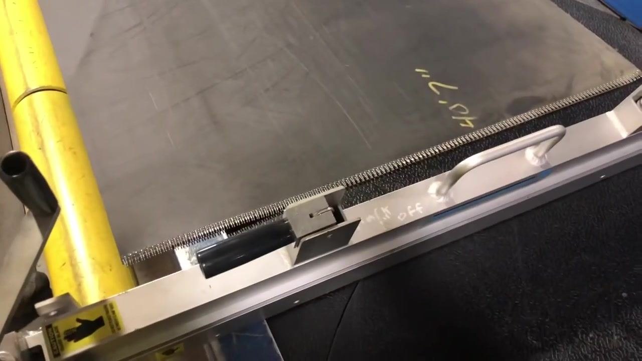 A worker in safety gear stands next to a conveyor belt with a mounted belt cutter. The cutter has black handles, a metal frame, and caution labels.