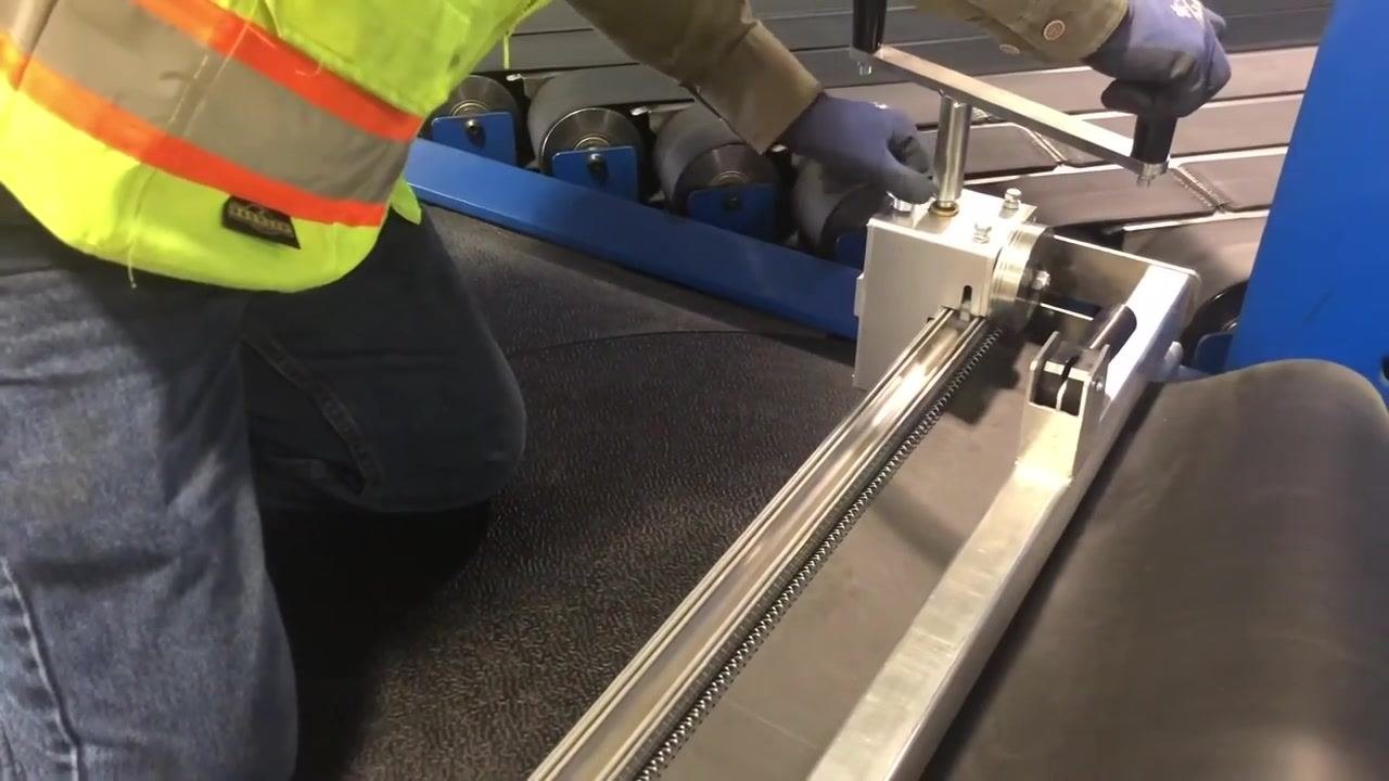 A worker in a high-visibility vest and gloves operates the lacing machine, moving the handle back and forth to lace the belt.