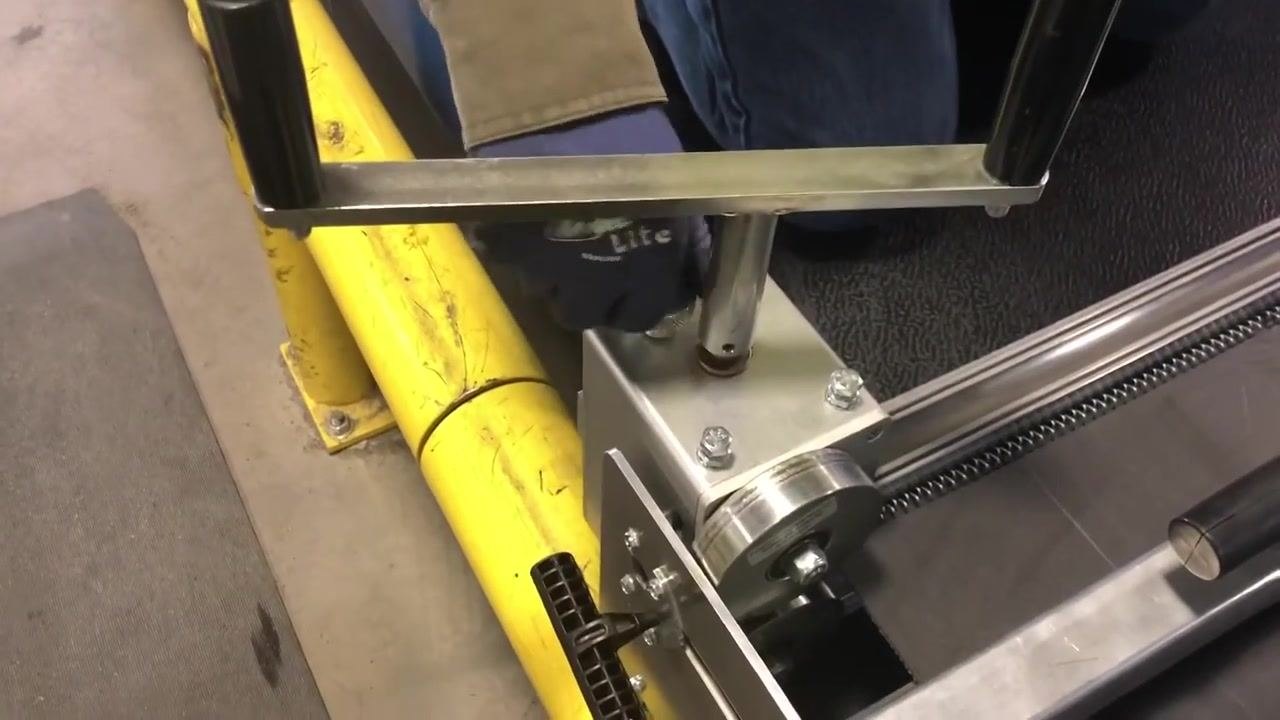 A worker in gloves kneels beside the lacing machine, preparing to inspect the nearly finished lacing. The environment includes a yellow safety barrier, a textured floor mat, and the metallic lacing machine with visible bolts and rollers.