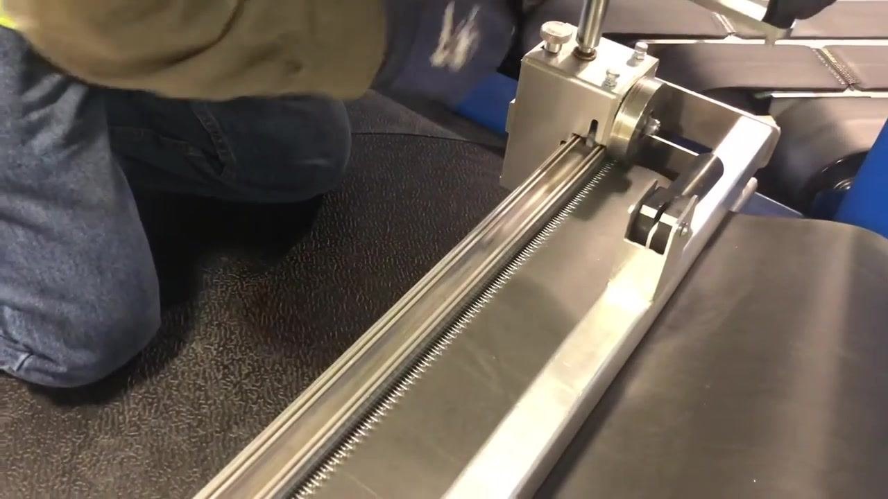 A gloved hand is running over a metallic lacing machine, with a black belt and visible hooks being pressed through the belt. The environment appears to be an industrial or workshop setting.