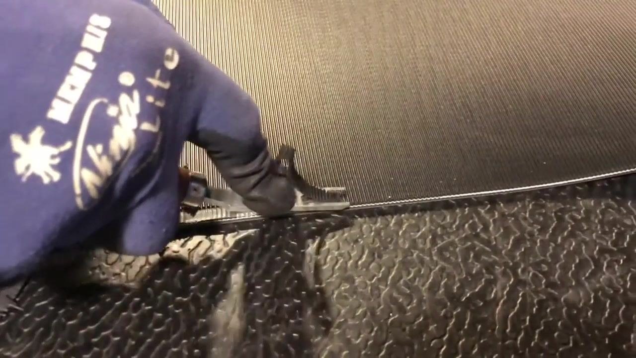 A gloved worker uses a skeeving tool to remove ribs from the edge of a rough-textured conveyor belt.