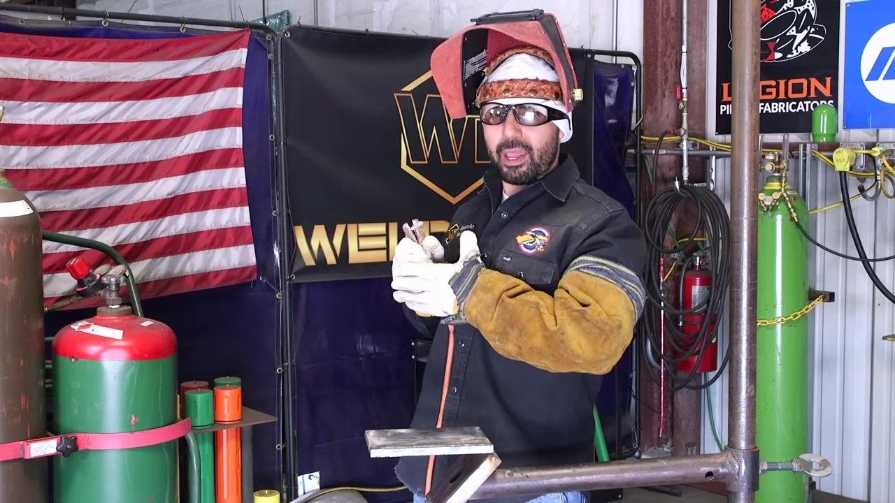 A welder in full safety gear stands upright with hands together, explaining posture, with welding equipment visible in the background.