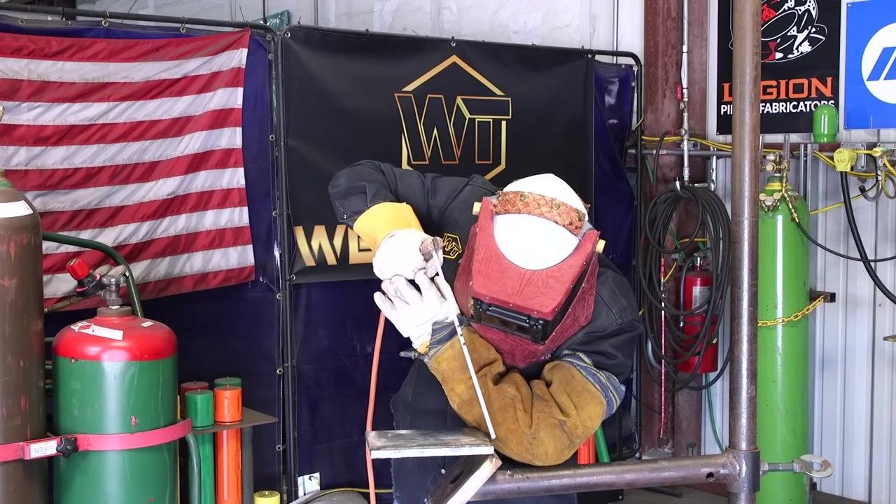 A welder is actively welding with the helmet down, focused on the workpiece, with visible arc and sparks.