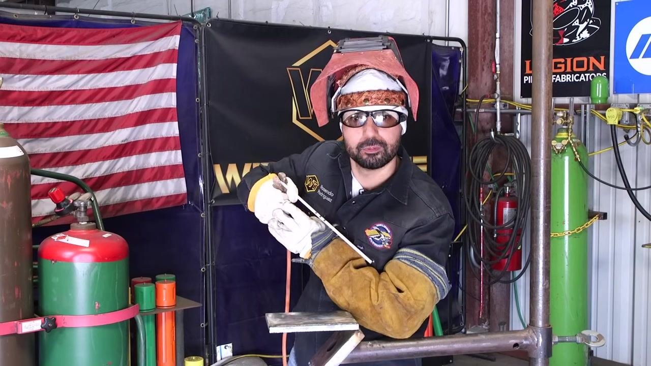 A bent-knee, forward-leaning posture is demonstrated, with the welding rod held above the workpiece, ready to weld.