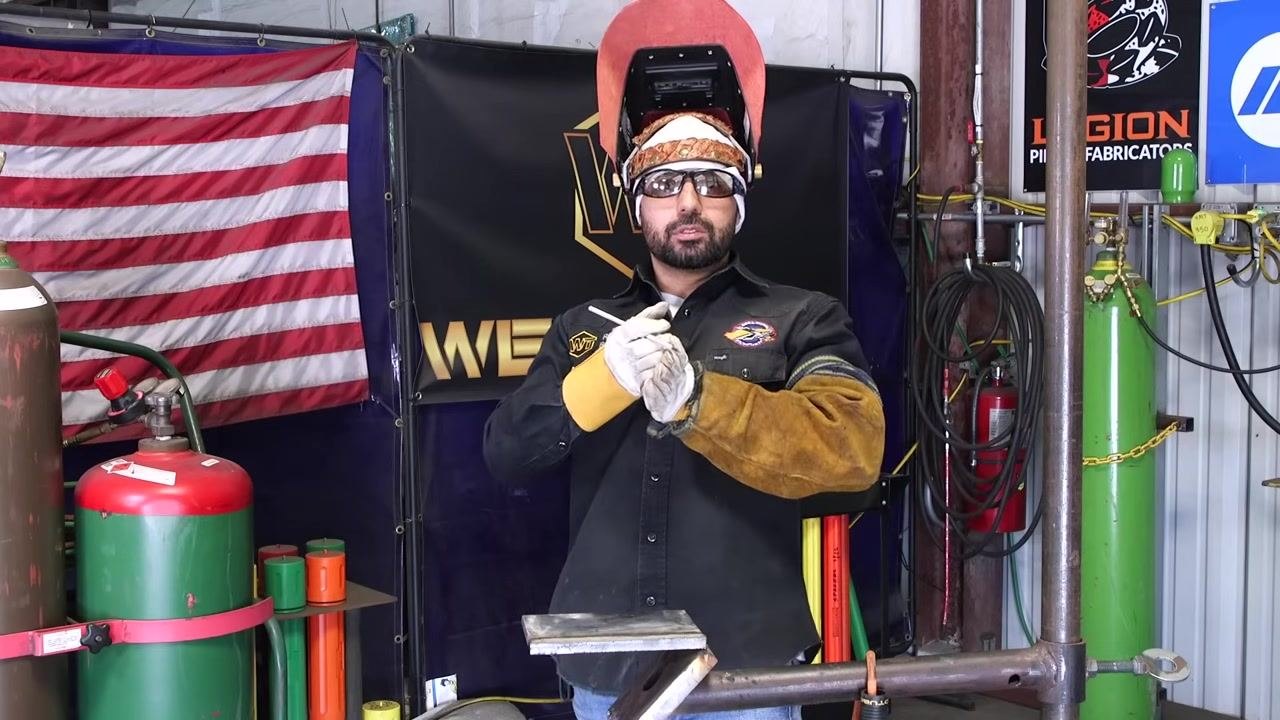 A welder stands upright, holding the rod and gesturing while explaining troubleshooting tips.