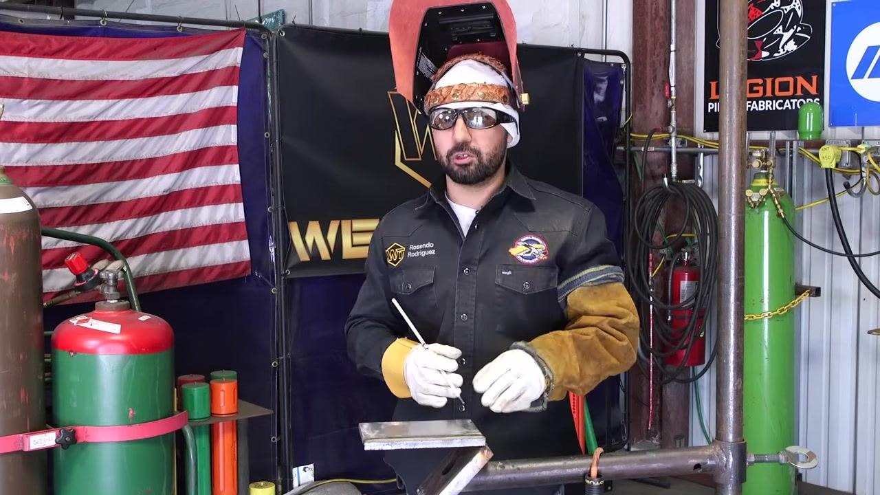 A welder holds a welding rod while wearing full safety gear, with an American flag, gas cylinders, and welding banners in the background.