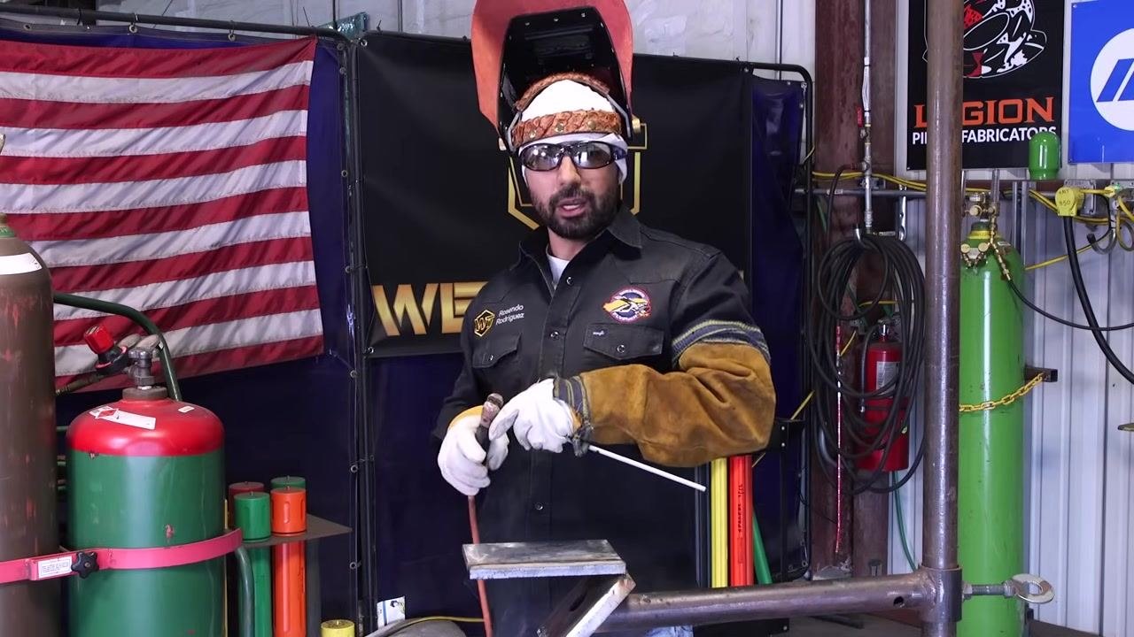 A welder holds a welding rod and stinger, demonstrating proper grip, with an American flag, gas cylinders, and welding equipment visible in the background.