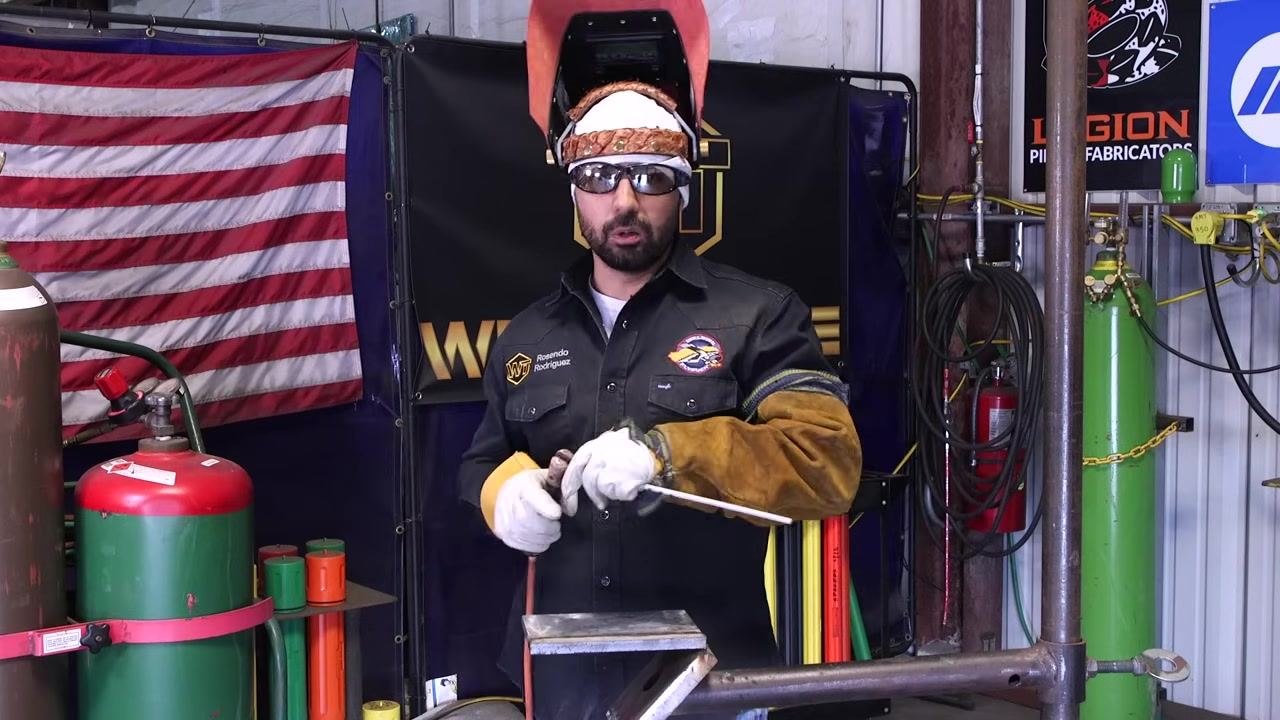 A welder in full gear holds a welding rod and torch, with safety equipment and gas cylinders visible in the background.