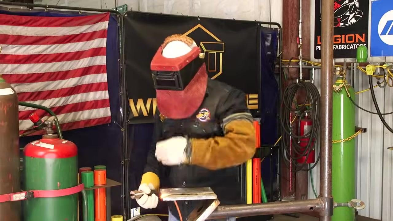 Instructor in welding gear, standing at the welding table, signaling the end of the demonstration. American flag, WT banner, gas cylinders, and welding equipment visible.